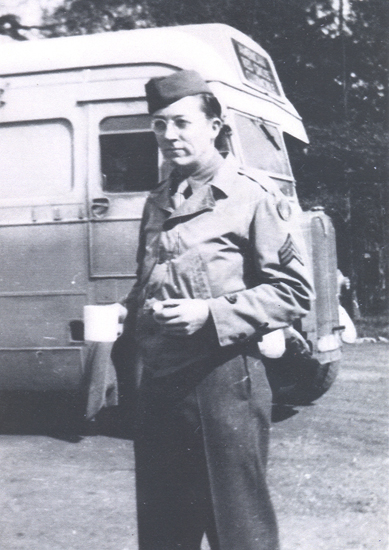 A man in uniform stands next to an old van, holding a cup. He wears glasses and has a backpack strapped over one shoulder.