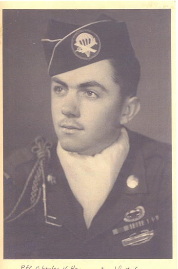 An aged, sepia-toned black and white photograph of a man in military attire, including a military beret adorned with an insignia, a tie, and a shoulder patch or emblem that could represent a specific military branch or country. The individual has a mustache.