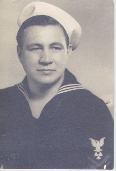 A sailor in uniform, standing and posing for a portrait. He has a serious expression, dressed in a navy uniform with a jacket and tie, and a white hat.
