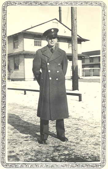 This is an old black-and-white photograph featuring a man dressed in military attire, standing on a snowy surface with a building and a utility pole visible in the background.