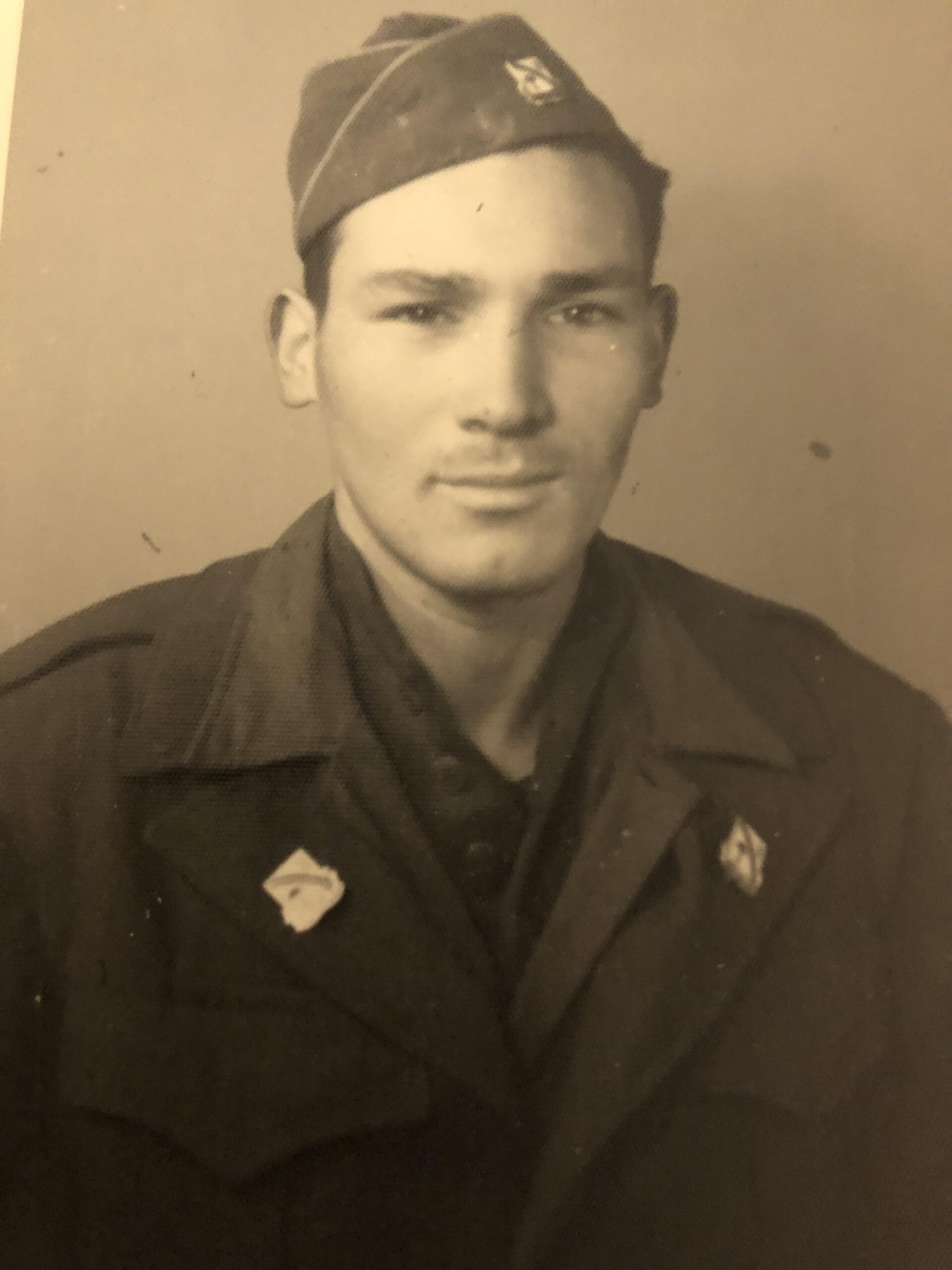 A vintage sepia-tonded photograph features a manwearing military attire, including a shirt with insignia and a tie. The person is facing forward and has short hair.