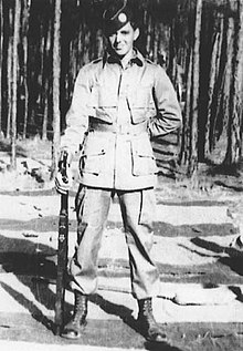 A man in military uniform holding rifle, standing outdoors in a wooded area with trees in background.