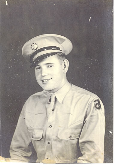 The image is a black-and-white photograph of a man wearing a military uniform. He has short hair and is smiling at the camera, looking directly into it.