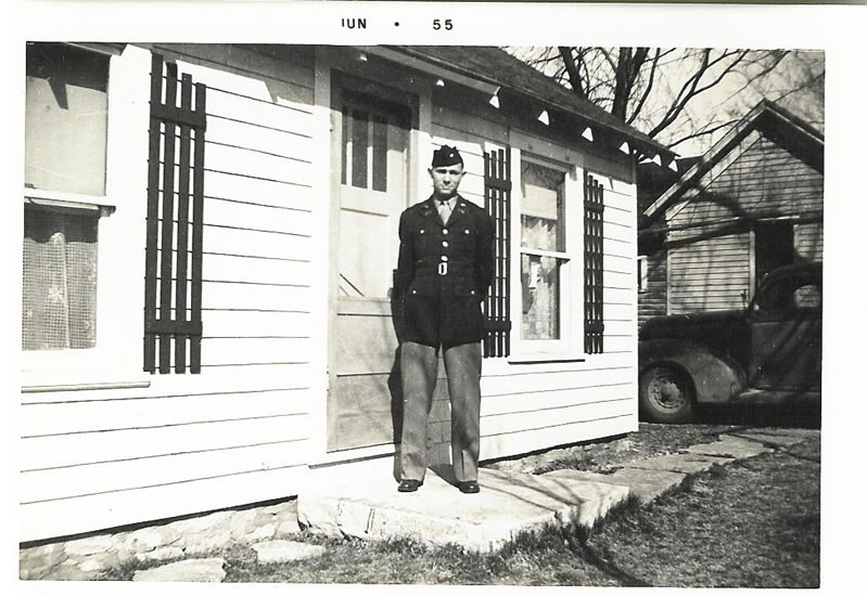 This is an old black-and-white photograph of a man standing in front of a house. They appear to be wearing a military uniform, and they are looking directly at the camera with a neutral expression. The house has a small porch, and there's a car parked by the house.