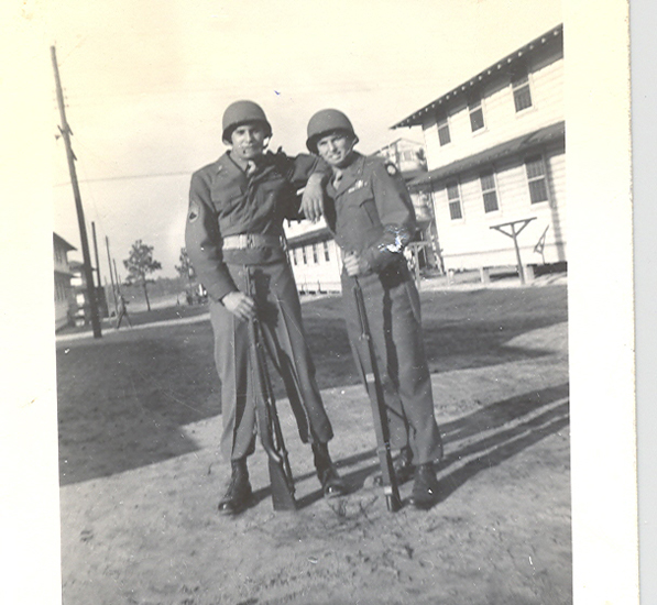 Two men in military uniforms, one leaning on the other, both standing outdoors with a vintage vibe, in front of an old structure.