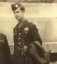 In black and white, an army man in uniform stands proudly. He is holding a folded jacket with one arm.