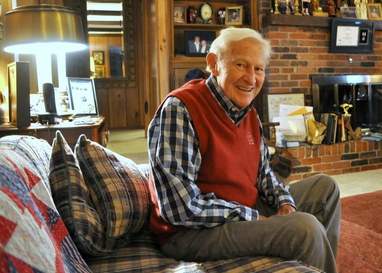 Older man seated indoors wearing a red sweater vest.