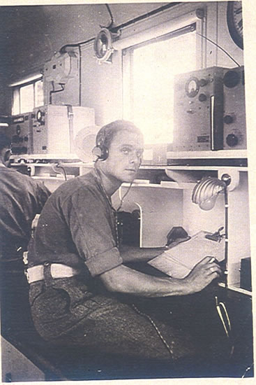 Man seated indoors at a desk with communications equipment.