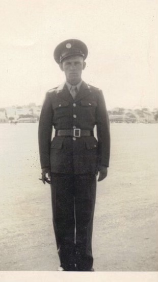 The image shows a man in military uniform standing. He is wearing a military cap, and his attire includes a tie.