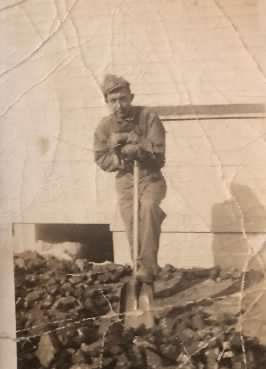 Serviceman standing outdoors in a military field uniform, leaning against a shovel.