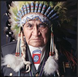 Native American elder wearing a feathered headdress while speaking into a microphone.