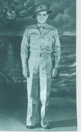 This is a black-and-white photo of a person standing, wearing a military dress uniform which includes a tie, belt, and tunic, along with cap. The individual appears to be posing for the photograph.