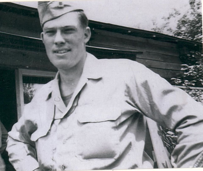 The image is of a man in military uniform standing outdoors. He is looking directly at the camera with a slight smile.