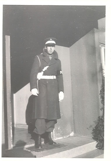 Serviceman standing in a doorway wearing a long dark uniform.