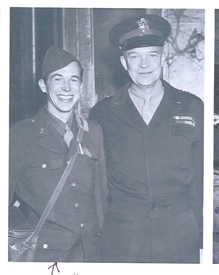This is a black and white photo featuring two men in miltary uniform. The man on the left is smiling at the camera. On his right side, there is Dwight D. Eisenhower. The background is indistinct, suggesting that the photo may have been taken at an event or during a formal occasion.
