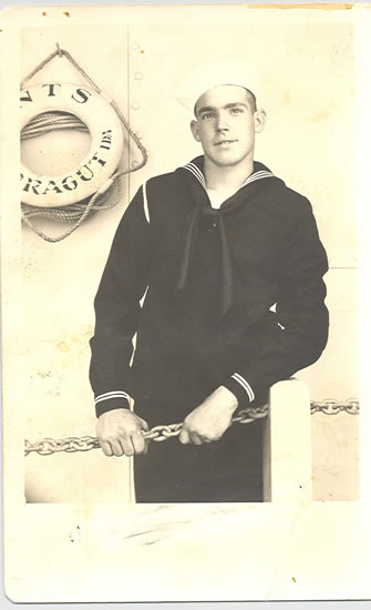 Sailor standing in a dark naval uniform beside a life preserver.