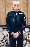 Older veteran standing indoors in a dark military jacket.