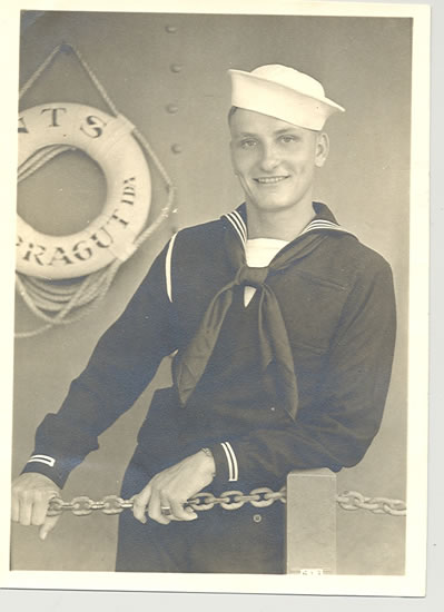 A sailor in dress uniform poses on a ship, standing proudly with his hands on a chain-style fence. He is smiling and looks directly at the camera.