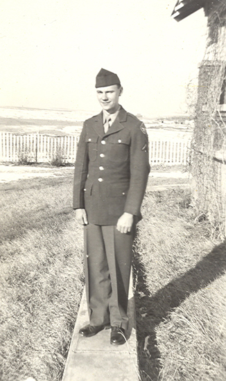 A soldier in military dress standing in front of a fence on grassy terrain. He has his hands by his side and wears a tie. The background shows a clear sky.