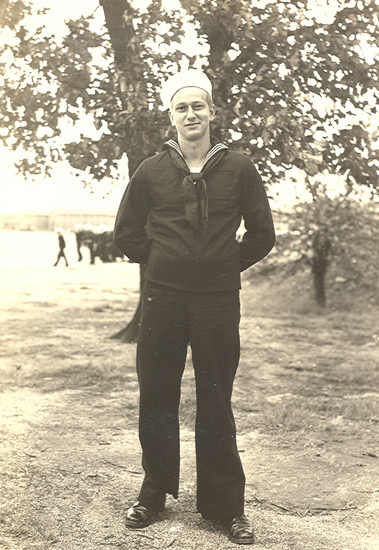 Sailor standing outdoors beneath a tree in a dark naval uniform.