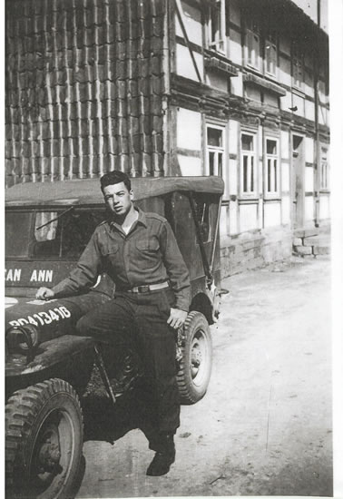 Serviceman seated outdoors beside a military jeep in military uniform.