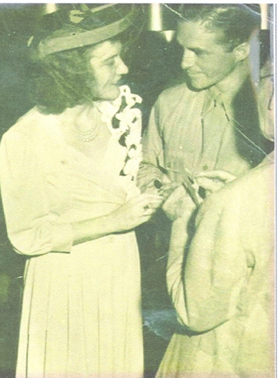 Two people standing near each other in what appears to be a social gathering, with one person wearing a tie and the other a hat. The setting looks like an indoor event, possibly a party or reception.