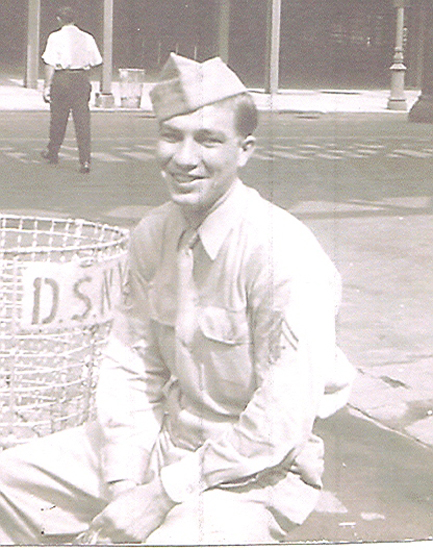 Smiling serviceman standing outdoors beside a handwritten sign.