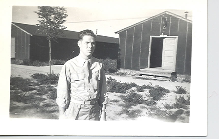 A black-and-white photograph features an individual standing in front of a building. The person appears to be wearing a uniform of some kind.