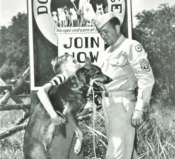 Soldier poses with a military dog beside a Dogs for Defense recruitment sign.