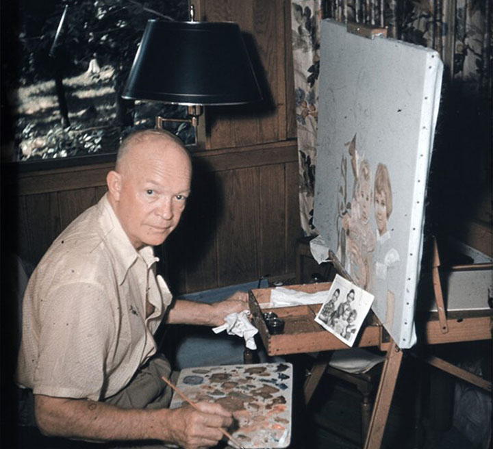 Dwight D. Eisenhower seated at an easel while painting indoors.