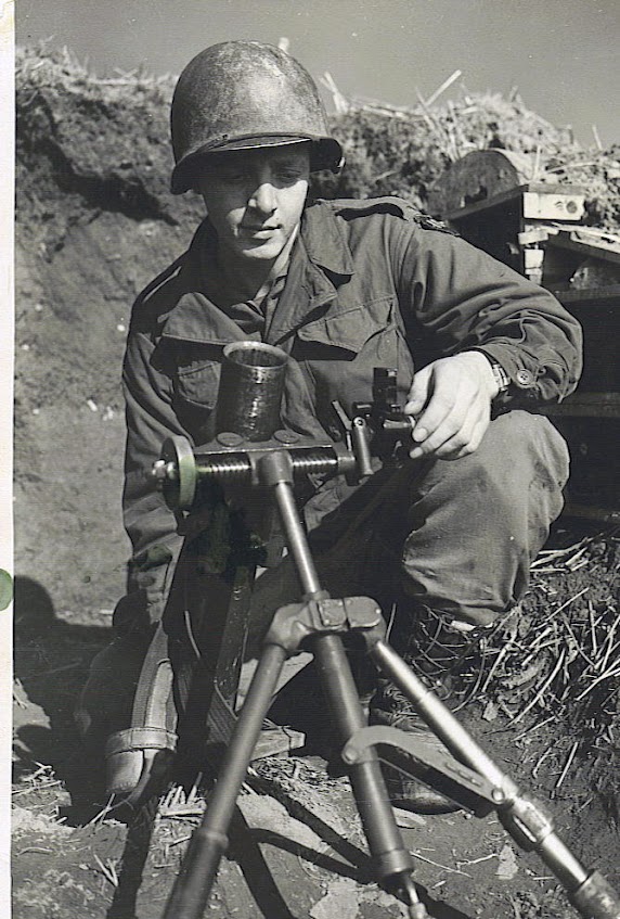 Soldier seated outdoors beside a mortar and other military equipment.