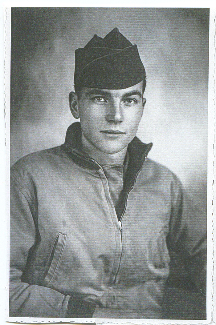 A man in military attire is seated and looking slightly off-camera with a slight smile. They are wearing a dark uniform and a cap. They are posing in front of a plain background.