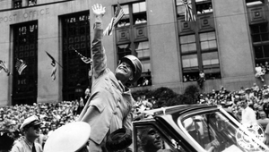 Black-and-white historical photograph of a smiling Eisenhower in a military uniform waving to a large crowd from an open-top car during a parade.