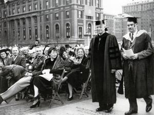 Historical black-and-white photograph of two men in academic caps and gowns walking together at an outdoor graduation ceremony, with a seated audience in the background.