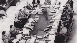 Black-and-white overhead photograph of participants seated around a large conference table covered with documents during an American Assembly meeting.